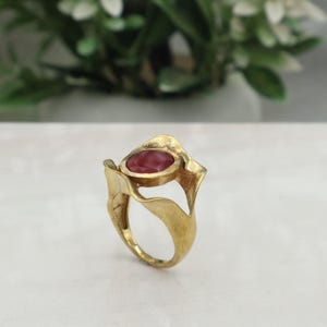 May include: A gold ring featuring a large, oval, red gemstone. The ring has an unusual, organic design with leaf-like features. The gemstone is set in a bezel, and the ring is shown on a white surface.