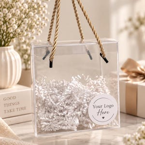 Pode incluir: Uma sacola de presente quadrada de plástico transparente com alças de corda dourada trançada. O interior é preenchido com papel picado branco. Um adesivo branco circular na frente apresenta o texto preto Your Logo Here com um coração.