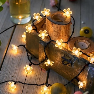 Op de afbeelding: Een lichtslinger met kleine, verlichte bloemvormige lampjes. De lampjes zijn bevestigd aan een zwarte draad en zijn gerangschikt rond een houten kist en kaarshouders. De bloemen hebben een zachte gele kleur, wat een warme sfeer creëert.