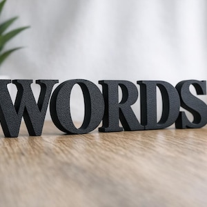 Könnte beinhalten: Schwarze, strukturierte Holzbuchstaben mit dem Wort „WORDS“ auf einer Holzoberfläche. Eine Topfpflanze, eine Kerze und ein Stapel Bücher befinden sich im Hintergrund. Die Buchstaben sind etwa 15 cm hoch.