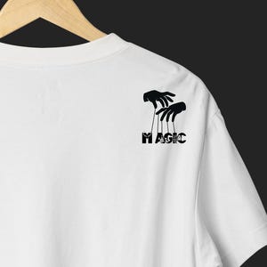 May include: White t-shirt with a black graphic of puppet hands and the word "MAGIC" in a stylized font. The design is located on the upper right shoulder area of the shirt.