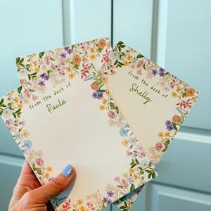 May include: Three floral stationery sheets are held in a hand. The sheets have a white background with a floral border in shades of pink, yellow, purple, and blue. The text "from the desk of Paula" and "from the desk of Shelly" are visible.