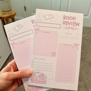 May include: Two pink book review sheets with a title, summary, and rating sections. The sheets have a decorative bow and book illustrations. The text "Book Review" is at the top of one sheet.