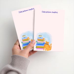 May include: Two pale pink rectangular notepads featuring the text Notes between chapters in dark blue. Each notepad displays a colorful illustration of a stack of books topped with a teacup and an orange tabby cat sitting beside the books.