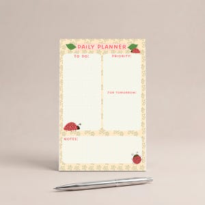 May include: A cream-colored daily planner with a ladybug and floral design. The planner has sections for to-do lists, priorities, and notes, with the text "DAILY PLANNER" at the top. A silver pen rests at the bottom.