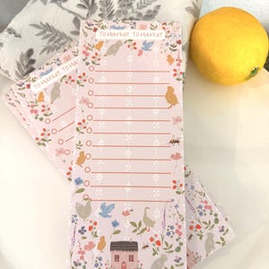 May include: Pink notepad with the text "To Market To Market" and a floral and bird design. The notepad has a list format with circles for checking off items. A lemon and a floral towel are in the background.