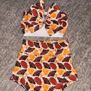May include: A two-piece baby outfit featuring a headband and high-waisted shorts. The set is patterned with a repeating design of orange and red shapes, and the word "TEXAS" in a red and green banner.