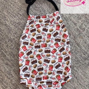 May include: A white baby romper with a repeating pattern of fast food items, including burgers, fries, and drinks. The romper has a black halter neck tie and the text "Sassy Sadie's Sewtique" in a pink logo.