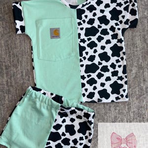 May include: A two-piece outfit featuring a mint green and black and white cow print design. The short-sleeved top has a mint green pocket with a Carhartt logo. The matching shorts have a mint green and cow print pattern. The outfit is designed for children.