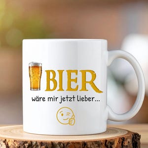 Könnte beinhalten: Weiße Keramik-Tasse mit dem Wort "BIER" in Gelb, einer Bierglas-Grafik und deutschem Text. Die Tasse steht auf einem Holzuntersetzer. Ein zwinkernder Smiley ist ebenfalls auf der Tasse.