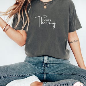 Könnte beinhalten: Dunkelgraues T-Shirt mit dem weißen Schriftzug "Books are my Therapy". Das Shirt wird mit blauen Jeans und weißen Turnschuhen getragen. Das Bild vermittelt einen entspannten, lässigen Eindruck, der auf bequeme Alltagskleidung hindeutet.