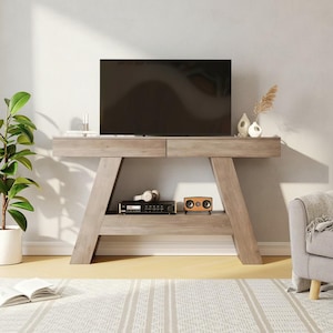 May include: A light gray wooden TV console with a large flat-screen TV on top. The console has two drawers and an open shelf with a stereo, headphones, and a small speaker. A potted plant and decorative vases are also visible.
