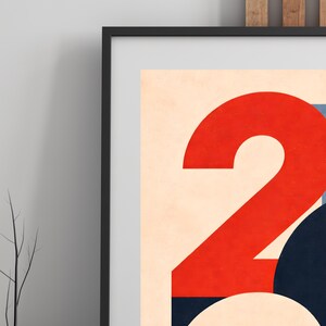 Pu&ograve; includere: Stampa artistica astratta con un grande numero "2" rosso su sfondo color crema. L'opera d'arte include forme geometriche blu e nere, il tutto incorniciato in un bordo nero. Lo stile ricorda il design moderno della met&agrave; del secolo.