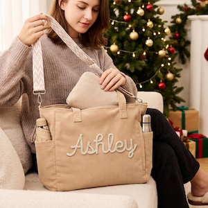 Op de afbeelding: Een beige corduroy tote bag met de naam Ashley in witte schrijfletters op de voorzijde. De tas heeft twee zijvakken, één met een kleine paraplu en de andere met een metalen fles, en is voorzien van een afneembare schouderband met patroon.