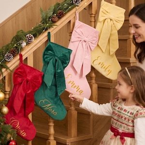 Könnte beinhalten: Vier Weihnachtssocken aus Samt in Rot, Grün, Rosa und Gelb, jeweils mit einer großen dekorativen Schleife und goldener Schreibschrift-Stickerei. Die Namen Olivia, Nora, Emma und Emily stehen auf den Socken, die an einer Holztreppe hängen.