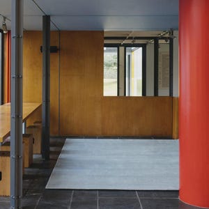 May include: Interior architectural shot featuring a modern design with a long wooden table and benches. The space includes a red pillar, wooden paneling, and a gray rug. Large windows offer a view of the outside.