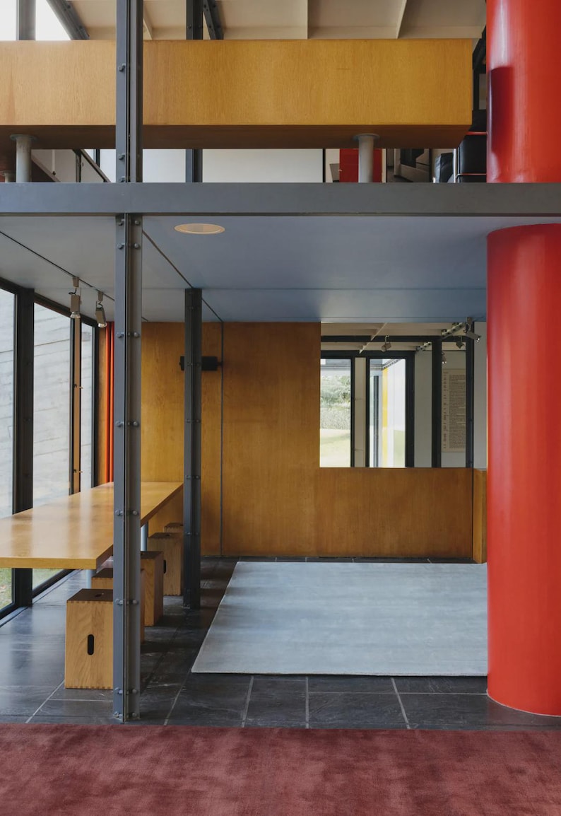 May include: Interior architectural shot featuring a modern design with a long wooden table and benches. The space includes a red pillar, wooden paneling, and a gray rug. Large windows offer a view of the outside.