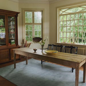 May include: A light-filled dining room with a long, weathered wooden table, a blue rug, and a wooden cabinet. The room has large windows overlooking greenery, and a black bench is placed against the windows.