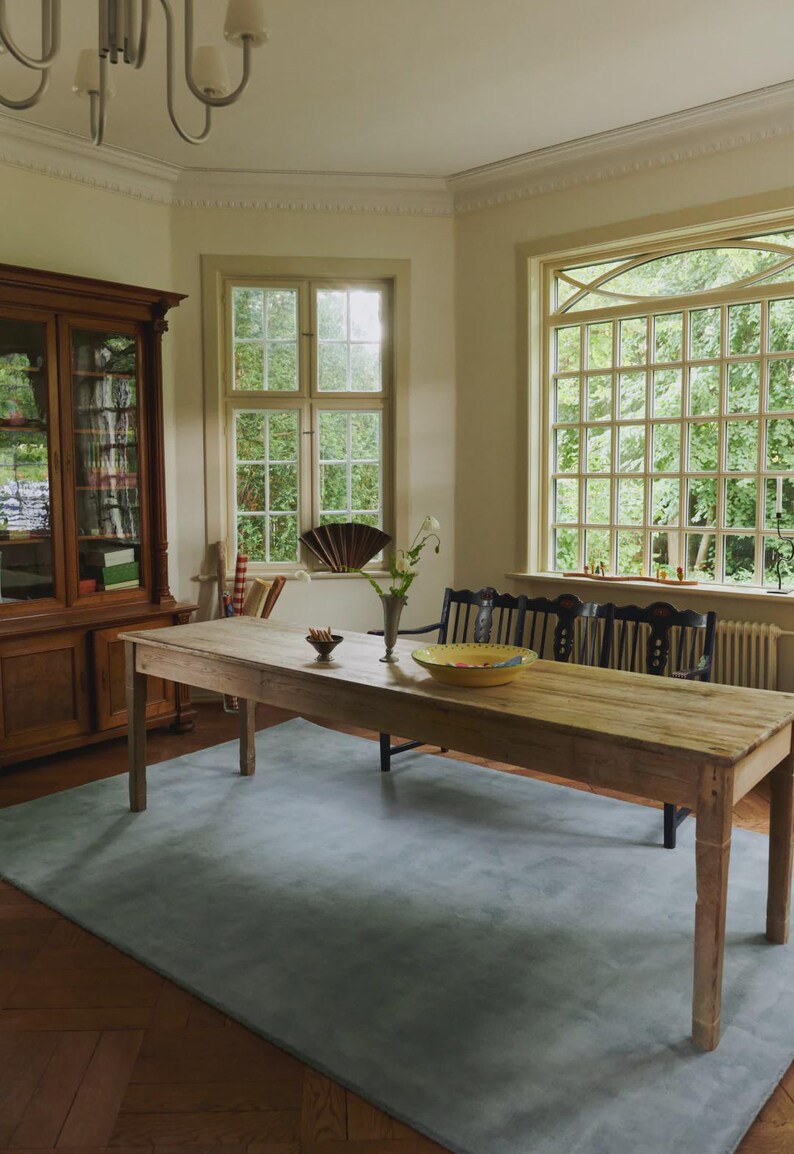 May include: A light-filled dining room with a long, weathered wooden table, a blue rug, and a wooden cabinet. The room has large windows overlooking greenery, and a black bench is placed against the windows.
