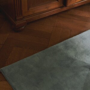 May include: A soft, gray rug with a plush texture, angled across the frame. The rug contrasts with the warm, brown tones of the wooden parquet flooring and a dark wooden cabinet in the background. The scene is lit with natural light.