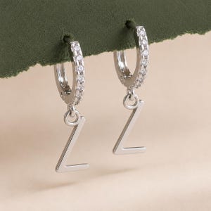May include: A pair of silver-toned hoop earrings featuring a row of small, clear, sparkling stones along the front of each hoop. Each earring has a dangling charm in the shape of the capital letter Z with a smooth, polished metallic finish.