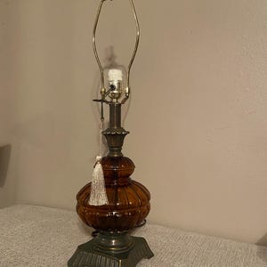 May include: A vintage table lamp with a brown glass body and a brass-colored metal frame. The lamp has a decorative tassel and a square base. The lamp is designed for a standard light bulb.