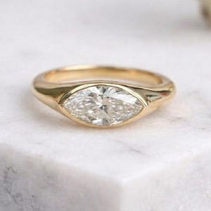 Op de afbeelding: Een gouden ring met een marquise-geslepen diamant. De diamant is gezet in een bezel-zetting en de band is glad en gepolijst. De ring wordt gepresenteerd op een wit marmeren oppervlak.
