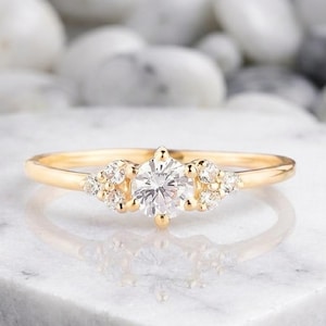 Op de afbeelding: Een gouden verlovingsring met een ronde, heldere middensteen en kleinere accentstenen aan weerszijden. De ring is gezet op een gouden band en wordt gepresenteerd op een wit marmeren oppervlak.
