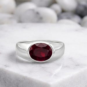 May include: A silver ring with a large, oval-cut, deep red gemstone. The ring has a smooth, polished band and a bezel setting that securely holds the gemstone. The ring is displayed on a white marble surface.