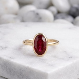 Könnte beinhalten: Ein goldener Ring mit einem großen, oval geschliffenen Rubin. Der Rubin hat eine tiefrote Farbe und ist in einem schlichten Goldband gefasst. Der Ring wird auf einer weißen Oberfläche präsentiert.