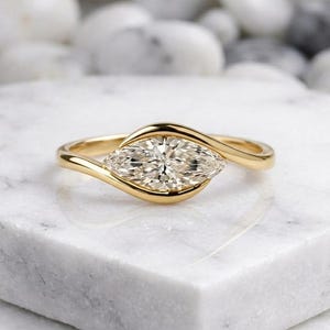 May include: A gold engagement ring featuring a marquise-cut diamond. The diamond is set in a swirling, bypass-style band. The ring is displayed on a white marble surface, with white stones in the background. The ring is a piece of fine jewellery.