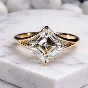 May include: A gold ring featuring a large, square-cut diamond. The diamond is set in a gold band with a delicate, open design. The ring is displayed on a white marble surface, showcasing its brilliance and clarity.
