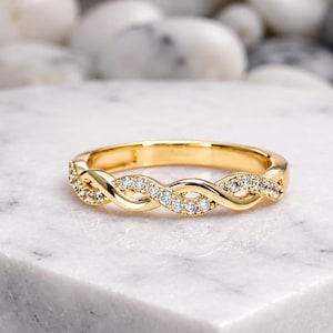 May include: A gold ring featuring a braided design, adorned with small, clear gemstones. The ring is presented on a white marble surface, highlighting its intricate details and elegant style. The ring's design is a continuous, interwoven pattern.