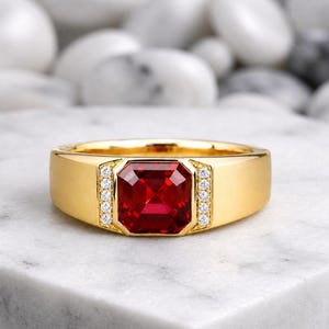 Könnte beinhalten: Ein goldener Ring mit einem großen, rechteckigen, roten Edelstein in der Mitte. Der Ring hat ein dickes Band und ist mit kleinen, klaren Edelsteinen auf beiden Seiten des zentralen Steins verziert. Der Ring wird auf einer weißen Marmoroberfläche präsentiert.