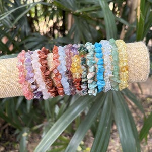 May include: A collection of colorful gemstone chip bracelets displayed on a tan fabric-covered cylinder. The bracelets feature various colors, including pink, purple, brown, blue, and yellow. The background is a lush green with leafy foliage.