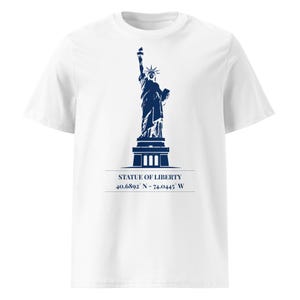 Può includere: T-shirt bianco con grafica blu navy della Statua della Libertà. Il design include il testo "STATUE OF LIBERTY" e le coordinate 40.6892° N - 74.0445° W. La maglietta è realizzata in un materiale morbido e confortevole.