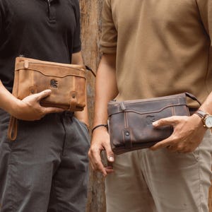 Pode incluir: Duas bolsas de mão de couro marrom com fecho de zíper e alça de pulso. As bolsas têm uma aparência texturizada e um logotipo na frente. Uma é marrom claro, a outra é marrom escuro.