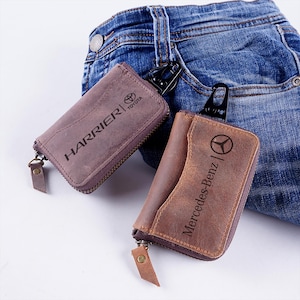 May include: Two leather key wallets, one brown and one purple, with zippers and metal clasps. The brown wallet is embossed with "Mercedes-Benz", and the purple wallet says "Harrier | Toyota". Both are resting on denim.