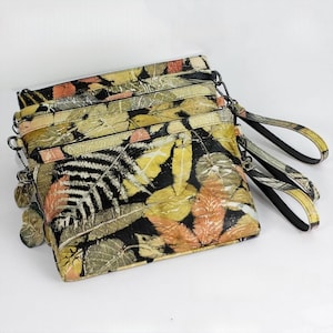 May include: A set of three leaf-patterned clutch bags. The bags feature a black background with yellow, orange, and green leaf designs. Each bag has a wrist strap and a silver-tone chain. The bags are stacked on top of each other.