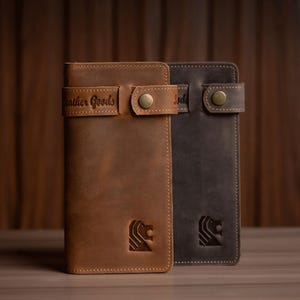 May include: Two leather wallets, one in tan and the other in charcoal, are displayed against a wooden backdrop. Each wallet has a strap closure with a brass button and a debossed lion logo. The tan wallet has the text "Leather Goods".