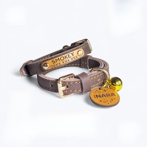 May include: Two brown leather cat collars with gold-colored hardware. One collar has the name "SMOKEY" and a phone number. The other collar has a gold bell and a tag that says "INARA".