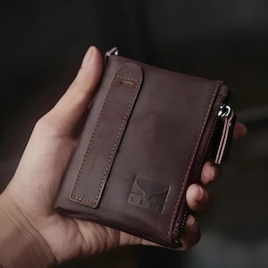May include: A brown leather wallet with a zip closure and a strap. The wallet has a logo on the front and is held in a hand. The wallet is approximately 10 cm wide and 10 cm tall.
