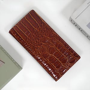 May include: A brown, rectangular wallet with a crocodile skin texture. The wallet is a rich, warm brown colour and appears to be made of leather. The wallet is lying flat on a white surface, with a book visible in the background.