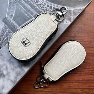 May include: Two white leather key fob cases with black trim and a silver-tone Honda logo. Each case has a zipper closure and a metal keyring attachment. The cases are on a wooden surface.