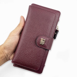 May include: A burgundy leather wallet with a textured surface. The wallet has a flap closure with a gold-colored metal accent that reads "ZHR". The wallet is held in a hand.