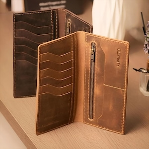 May include: Open, brown leather wallet with multiple card slots and a zippered compartment. The wallet has a distressed finish and the brand name "FABRIZZIO" is visible. Another similar wallet is in the background.