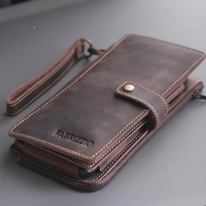 Puede incluir: Cartera de cuero marrón con correa de muñeca. La cartera tiene cierre de botón a presión y cremallera. La palabra "FABRIZZIO" está grabada en la parte delantera. Costuras blancas y cuero envejecido.