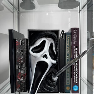Könnte beinhalten: Ein schwarz-weißer Scream-Masken-Buchstütze mit einem silbernen Messer, das Bücher hält. Zu den Büchern gehören "Haunting Adeline" und "The Psilocybin Mushroom Bible". Die Buchstütze wird in einer Glasvitrine ausgestellt.