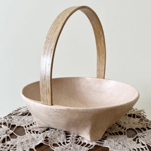 May include: A light-colored wooden basket with a curved handle. The basket has a square base and rounded bowl shape. The handle is arched and attached to the sides of the basket. The basket is sitting on a lace doily.