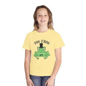 Könnte beinhalten: Ein hellgelbes T-Shirt mit einer Cartoon-Frosch, der einen Zylinder und eine Fliege trägt. Der Text "POG FROG" ist über dem Frosch aufgedruckt. Das T-Shirt wird von einer Person getragen.